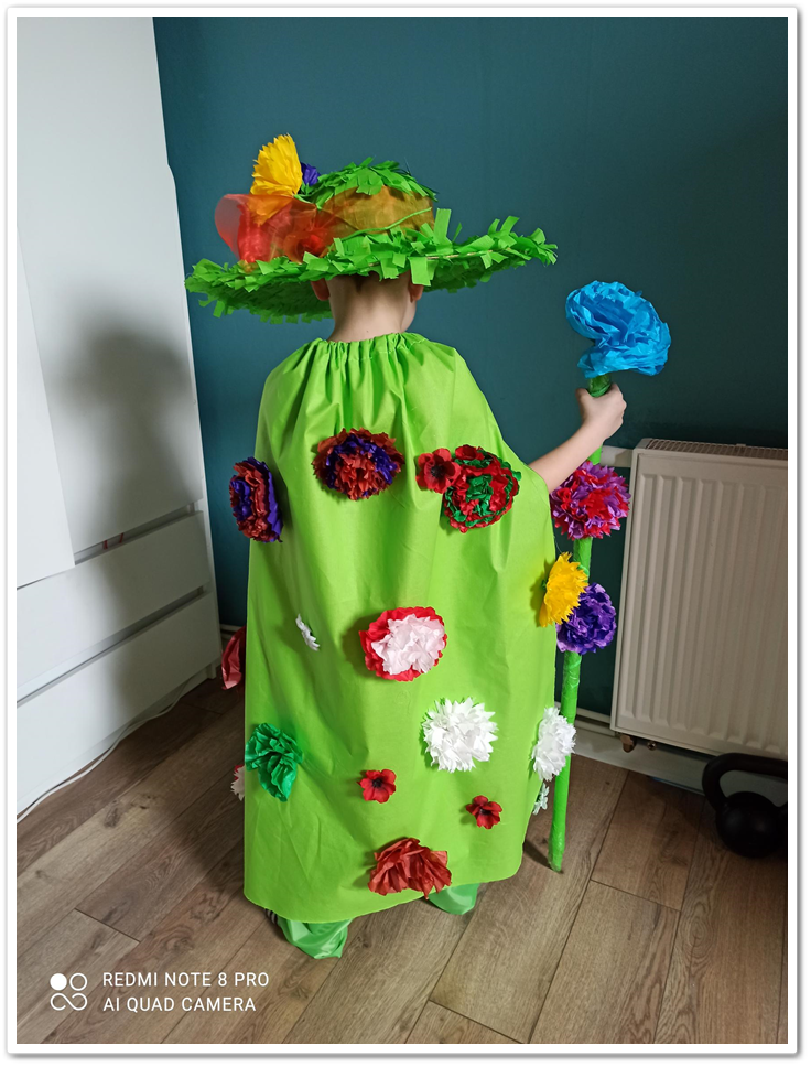 Chłopiec w przebraniu Pana Wiosny. Peleryna i kapelusz w kolorze zielonym, ozdobione kolorowymi kwiatami.