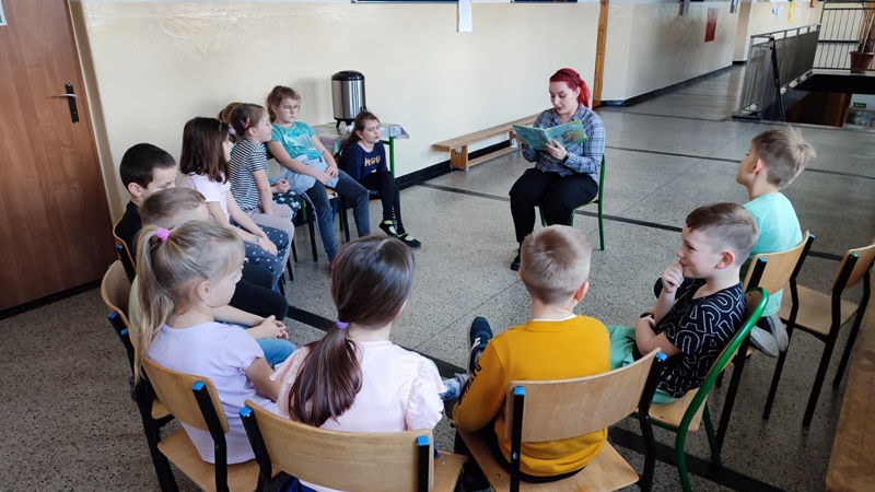 Dzieci siedzą w półkręgu, pośrodku bibliotekarka czytająca książkę