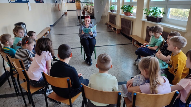 Dzieci siedzą w półkręgu, pośrodku bibliotekarka czytająca książkę
